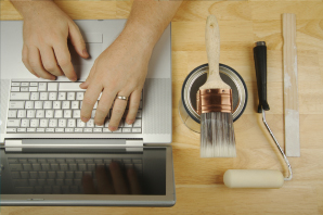 Picture of a hand typing on a laptop with a paintbrush, paint roller, and wooden paint stirrer